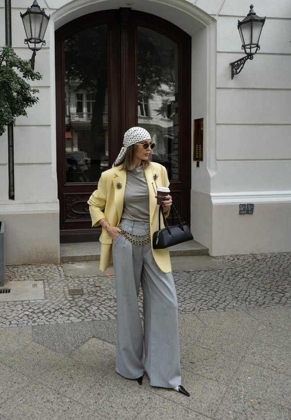 Femme en blazer jaune clair et pantalon gris à jambes larges debout sur le trottoir de la ville tenant un café et un sac à main noir.