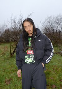 Doudoune noire avec capuche, rayures blanches sur les manches ; t-shirt graphique vert et noir ; pantalon noir ; cadre naturel en plein air.