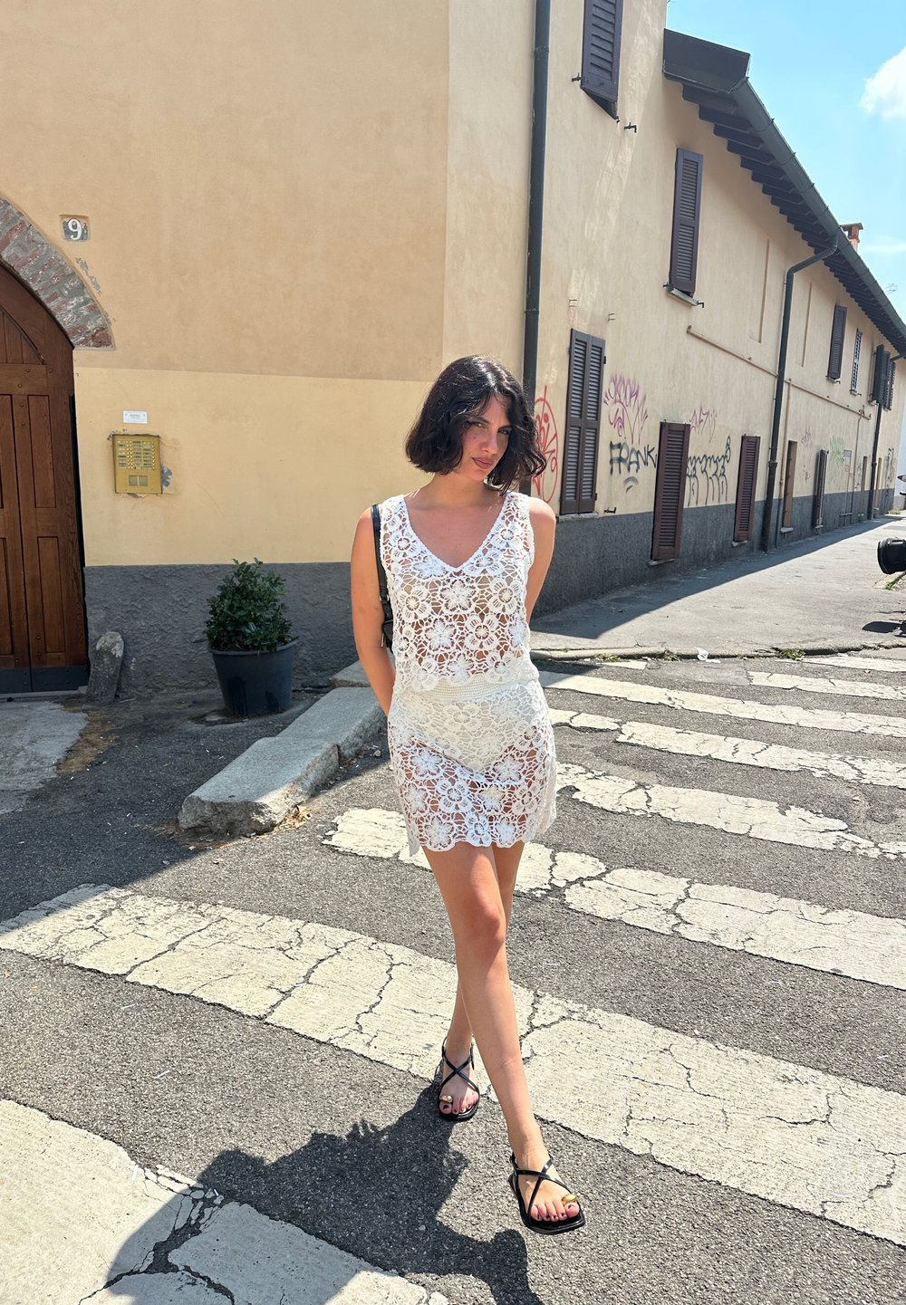 Abito di pizzo bianco con scollatura a V e vita aderente, caratterizzato da un design a gonna a strati. La modella indossa sandali neri con cinturini su un attraversamento pedonale.