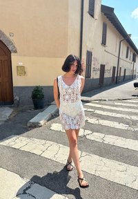 Abito di pizzo bianco con scollatura a V e vita aderente, caratterizzato da un design a gonna a strati. La modella indossa sandali neri con cinturini su un attraversamento pedonale.