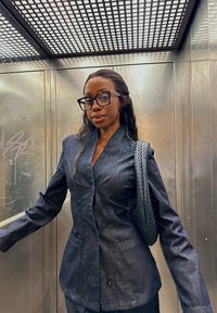 Dunkelblauer Denim-Blazer mit strukturiertem Design, zwei Vordertaschen und glatter Textur. Kombiniert mit einer Tasche mit geflochtenem Riemen. Moderner, taillierter Schnitt.