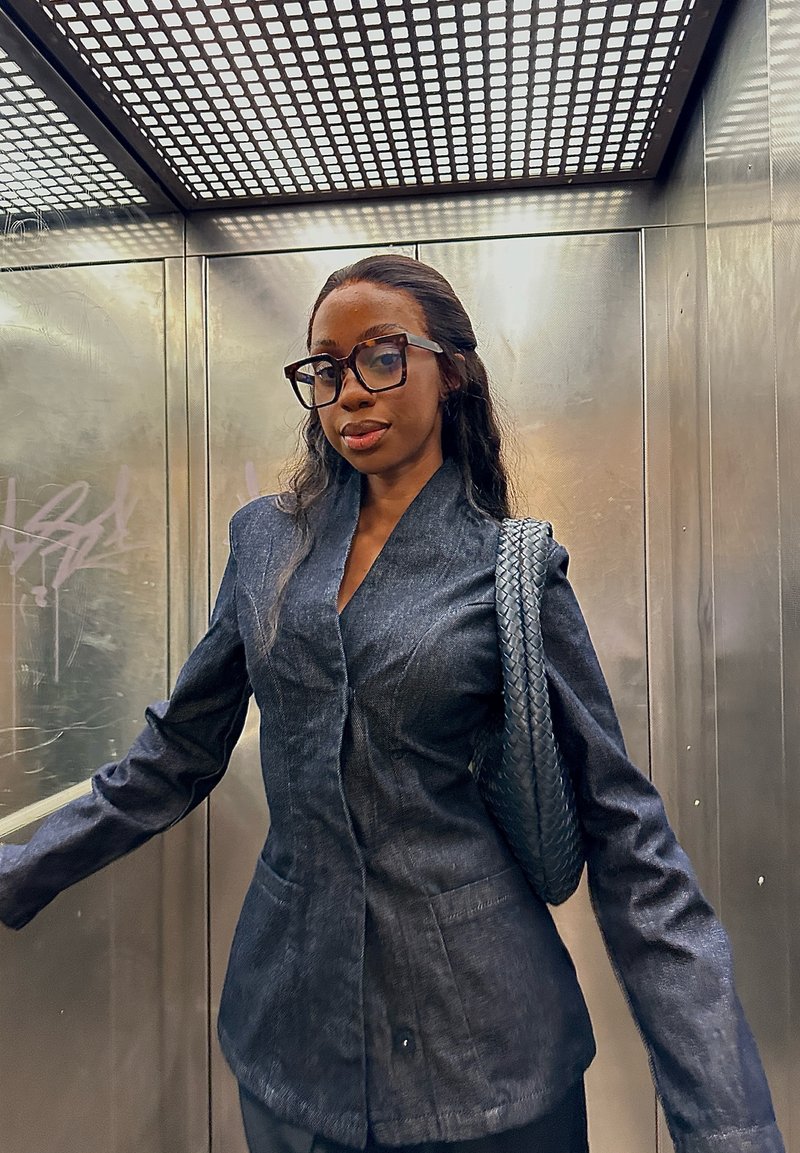 Dunkelblauer Denim-Blazer mit strukturiertem Design, zwei Vordertaschen und glatter Textur. Kombiniert mit einer Tasche mit geflochtenem Riemen. Moderner, taillierter Schnitt.