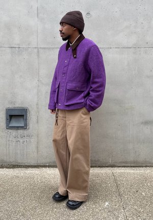 Hombre de pie junto a una pared de concreto, vistiendo una chaqueta morada, un gorro marrón, pantalones anchos beige y zapatos negros, con las manos en los bolsillos.