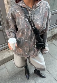 Person in einem camouflagierten Zip-Hoodie, grauen Shorts, schwarzen spitzen Stiefeln, silberner Kreuzkette und mit einer weißen Kaffeetasse im Freien.