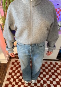 Personne portant un sweat-shirt gris à fermeture éclair et un jean large bleu clair, debout sur un tapis à carreaux marron et blanc à l'intérieur.