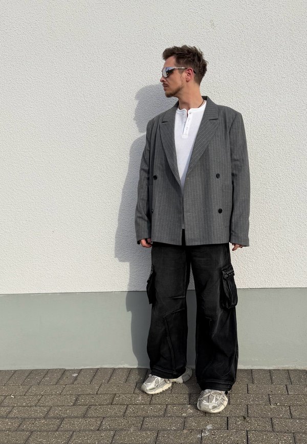 Mies, jolla on päällään harmaa raitainen bleiseri, valkoinen henley-paita, mustat cargo-housut ja lenkkarit, seisoo valkoista seinää vasten ja katsoo sivulle.