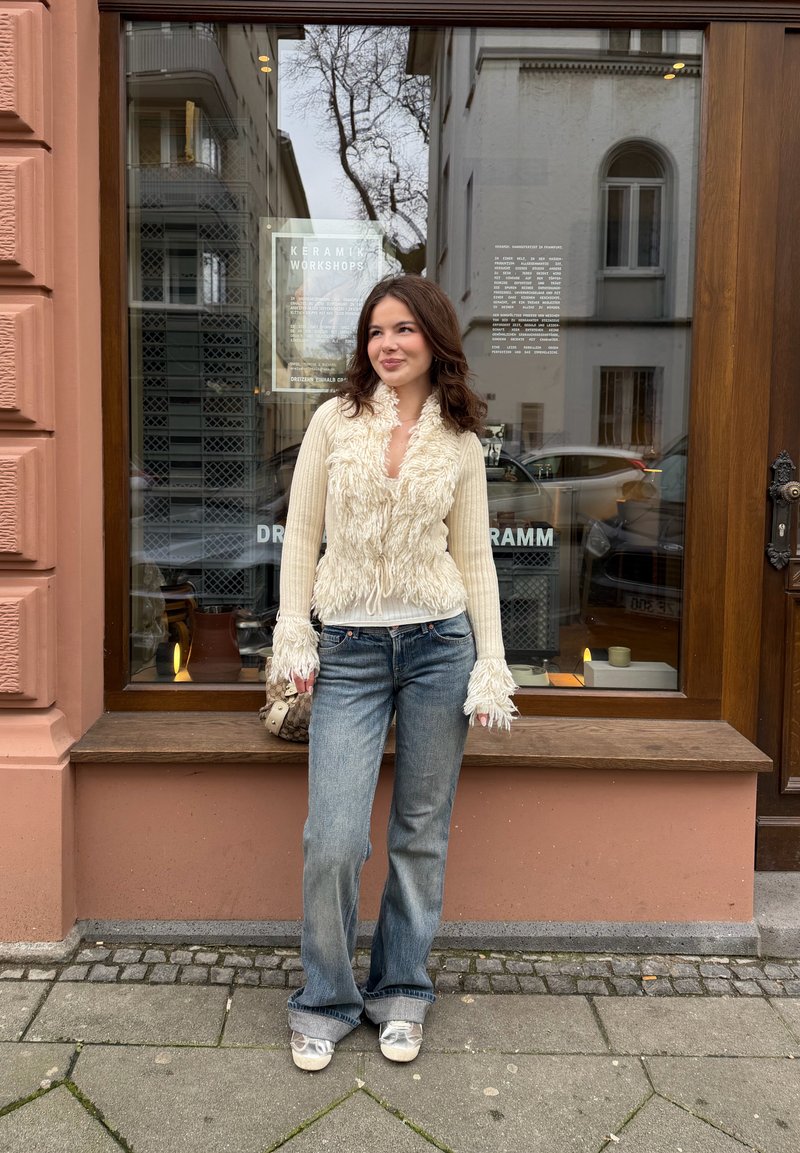 Jeune femme aux cheveux bruns ondulés portant un pull crème texturé et un jean ample, debout sur un trottoir devant une vitrine de magasin.
