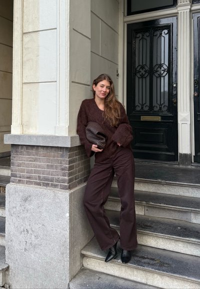 Suéter marrón peludo, pantalones anchos oscuros, zapatos de tacón negro puntiagudos, sosteniendo un bolso marrón texturado, apoyada contra un pilar de piedra cerca de puertas negras.