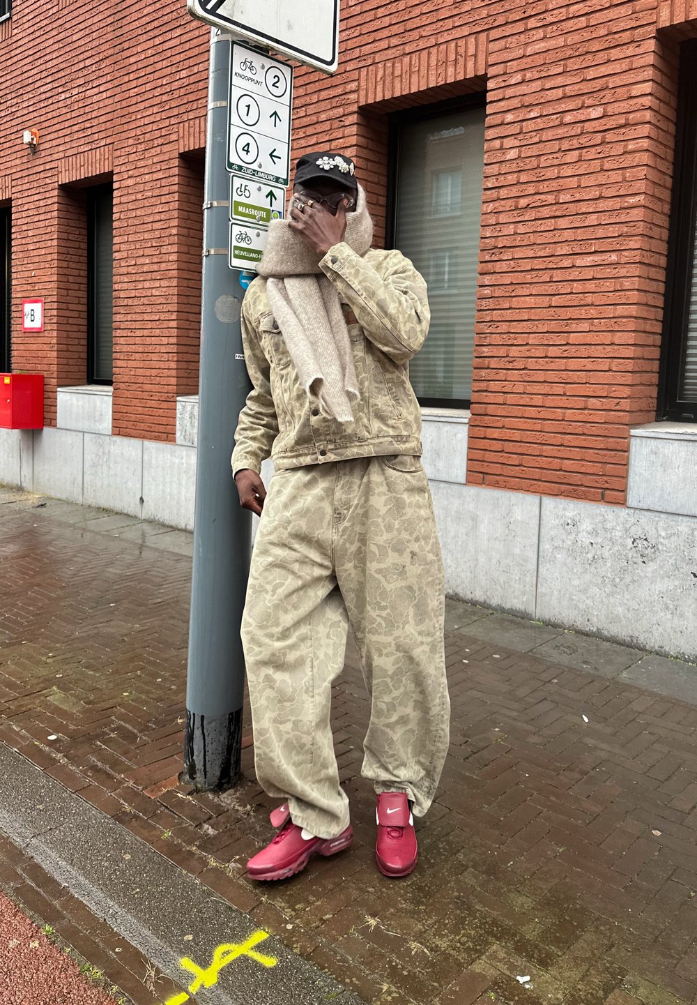 Μπεζ καπιτονέ μπουφάν με επένδυση και ταιριαστά φαρδιά παντελόνια, ανοιχτό καφέ κασκόλ, ροζ αθλητικά παπούτσια, στέκεται δίπλα σε στύλο με πινακίδες κυκλοφορίας μπροστά από τούβλινο τοίχο.