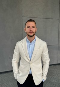 Világos bézs blézer egy világoskék ing felett, mindkettő texturált anyagból készült. Az egyén keze a zsebében áll, szürke háttér előtt.