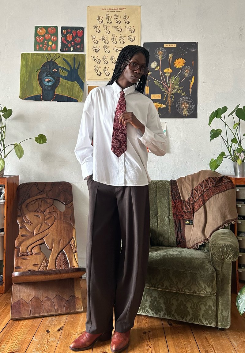 Personne ajustant une cravate rouge à motifs cachemire, portant une chemise blanche et un pantalon marron, debout dans une pièce avec des affiches botaniques et de langue des signes et une chaise en bois sculpté.