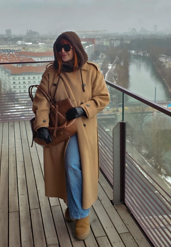 Femme portant un long manteau camel, une capuche, des lunettes de soleil et des gants, tenant un sac marron sur un balcon surplombant une rivière et une ville par une journée nuageuse.