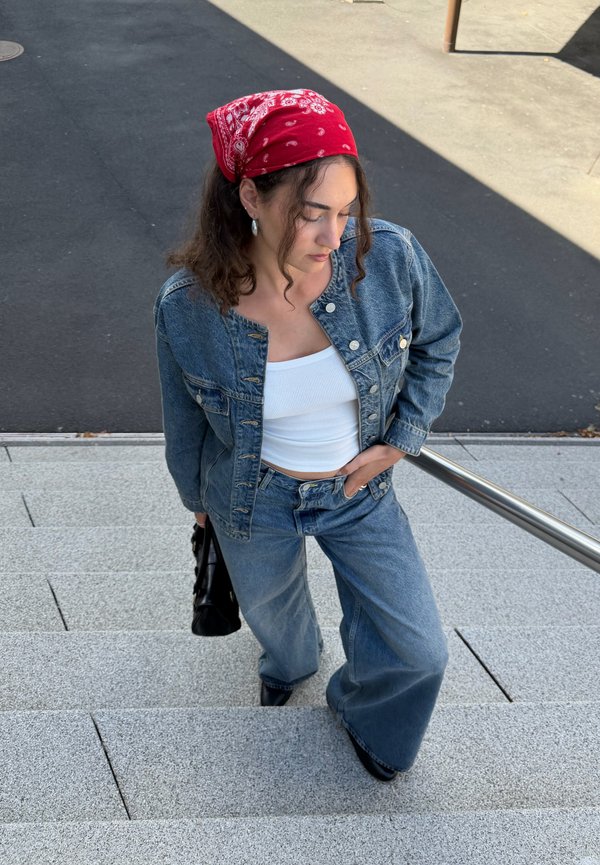 Mujer con bandana roja, chaqueta vaquera y vaqueros de pierna ancha subiendo unas escaleras al aire libre, llevando un bolso negro y mirando hacia abajo.
