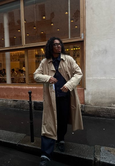 Hombre con pelo rizado y gafas, lleva un abrigo beige tipo gabardina sobre ropa oscura, y está de pie en una calle mojada cerca de la ventana de un café.