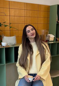 Asmuo su ilgais plaukais, dėvintis geltoną paltą ir kreminį megztinį, laiko kavos puodelį su latte meno piešiniu, sėdi prie žalių lentynų ir medinių sienų.