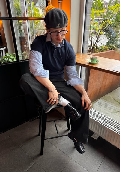Una persona está sentada en un taburete negro, vistiendo una camiseta polo azul marino, con mangas a rayas, pantalones negros y zapatos negros brillantes. Una taza de café verde está cerca.