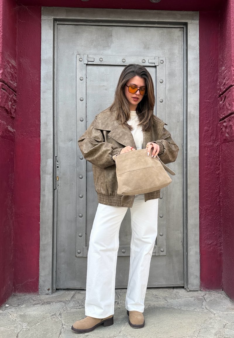 Frau trägt übergroße braune Jacke, weiße Hosen, hellbraune Stiefel und orange getönte Sonnenbrille, hält eine hellbraune Wildledertasche vor einer Metal tür.
