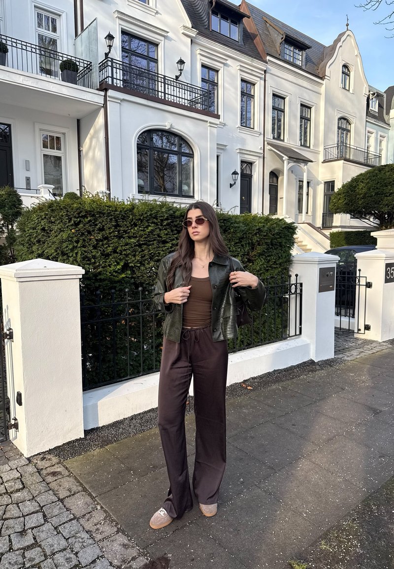 Jeune femme portant des lunettes de soleil noires, une veste verte, un haut marron et un pantalon ample foncé, debout sur un trottoir devant une maison de ville blanche et une haie.