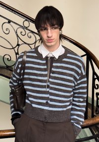 Jeune personne aux cheveux courts et foncés portant des lunettes, un cardigan rayé, une chemise blanche à col et un sac à bandoulière marron, debout près d'un escalier décoratif.