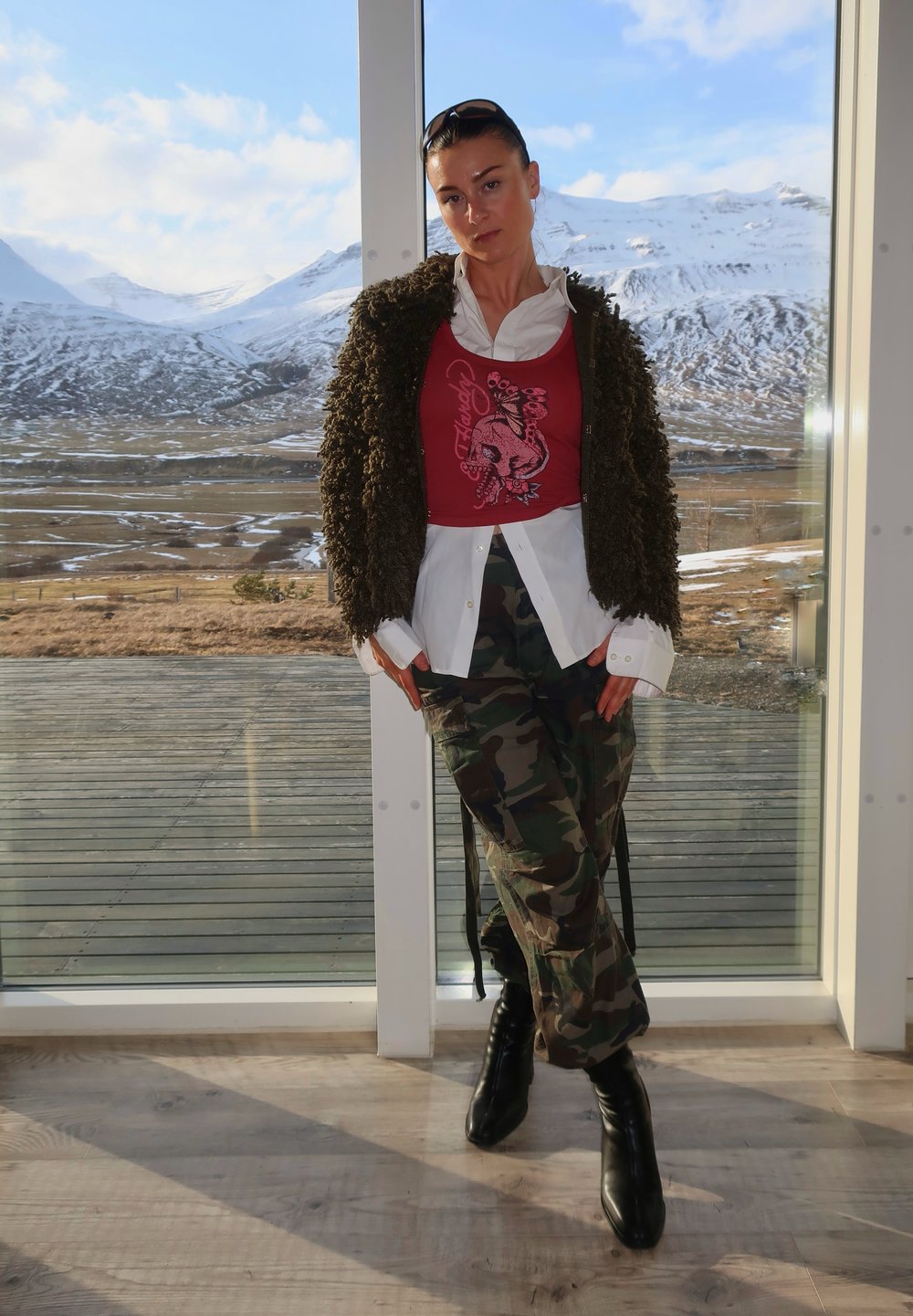 Um casaco castanho felpudo sobre um top cropped gráfico vermelho, uma camisa branca de botões e calças camufladas. Botas de salto alto pretas completam o visual.