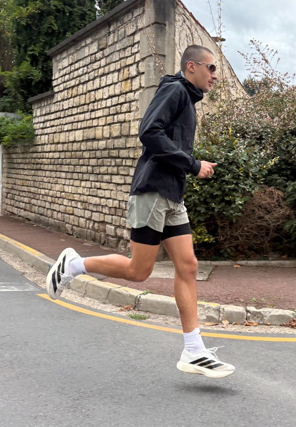 Un hombre con gafas de sol, una chaqueta negra y pantalones cortos de correr trota por una carretera junto a un muro de piedra y unos arbustos.