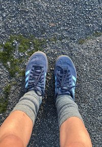 Blue suede trainers with light blue stripes, matched with grey ribbed socks. Feet placed on a rough, dark surface.