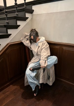 Mujer con abrigo beige y jeans azul claro sentada en un banco de madera bajo una escalera oscura, ajustándose un zapato de tacón negro abierto en los dedos.