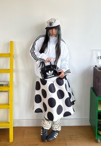 Bílá bunda s černými pruhy, sukně s puntíky, vzorované kalhoty, černé tenisky a lesklá černá kabelka s logem Adidas. Vertikálně pruhovaná čepice.
