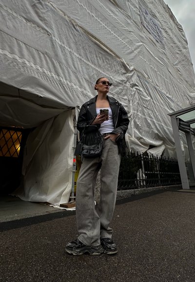 Chaqueta de cuero negra con flecos, camiseta blanca sin mangas, pantalones de pierna ancha grises y zapatillas negras. De pie al aire libre cerca de un sitio de construcción.