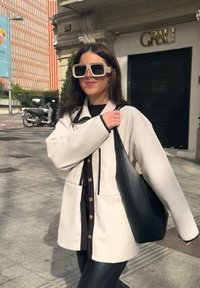 Υπέρμετρη κρέμα ζακέτα με μαύρες λεπτομέρειες και χρυσά κουμπιά, συνδυασμένη με μαύρη δερμάτινη τσάντα, φορεμένη πάνω από μαύρο μπλουζάκι και κολάν.