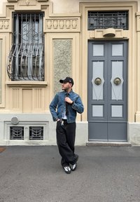 Jeansová bunda, šedé tričko, černé široké kalhoty a tenisky. V pozadí je zdobená budova se šedými dveřmi a dekorativními okenními mřížemi.