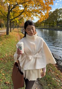 Witte, gestructureerde cape met een hoge kraag en strik op de taille. Model houdt een Starbucks-kopje vast, gekleed in een lichte rok en een bruine tas in een herfstpark.