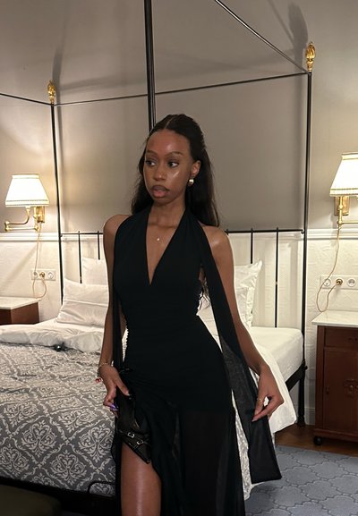 Mujer con un vestido negro de cuello halter de pie junto a una cama con una colcha de patrón gris y blanco en una habitación con dos lámparas.