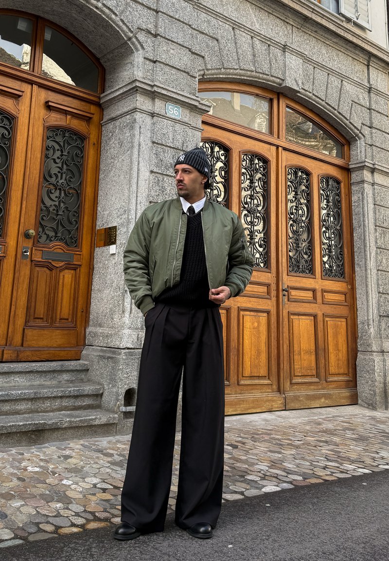 Homme portant une veste bomber verte, un pull noir, un pantalon large noir et un bonnet tricoté, debout sur une rue pavée devant un bâtiment en pierre avec des portes en bois.
