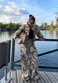 Kurtka w kamuflażowym wzorze i spodnie o szerokich nogawkach z teksturowanej tkaniny, w połączeniu z beżową torebką, na tle nadbrzeża.