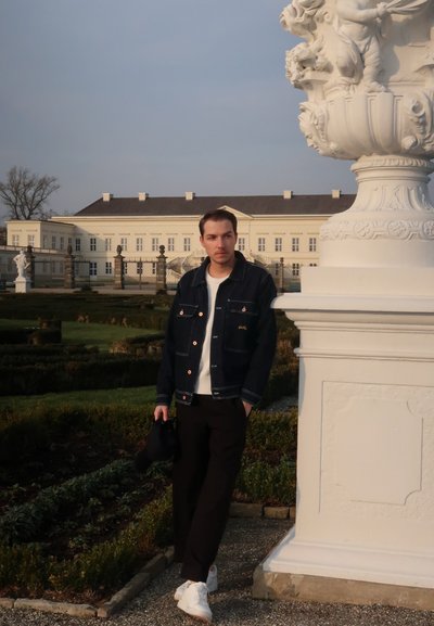 Chaqueta de denim con costuras en contraste sobre un suéter blanco, pantalones negros y zapatillas blancas; posando junto a una columna de piedra decorativa.