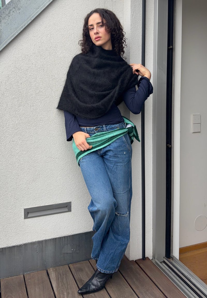 Woman with curly hair leans against wall wearing black shawl, navy long-sleeve top, blue jeans, black boots, and holding green striped sweater.