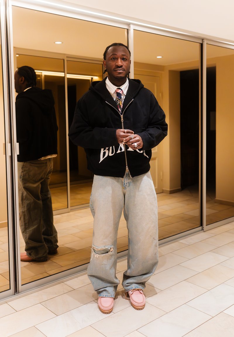 Άνδρας που στέκεται σε εσωτερικό χώρο φορώντας άνετο ξεσκισμένο τζιν, μαύρη ζακέτα με φερμουάρ πάνω από πουκάμισο και γραβάτα, ροζ παπούτσια, με καθρέφτη τοίχο πίσω του.