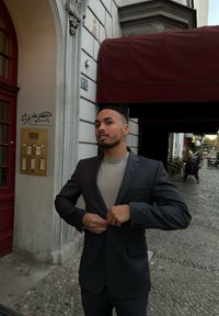 Ciemnoszara, strukturalna marynarka noszona na jasnoszarej koszuli. Osoba poprawia marynarkę przed budynkiem z czerwonymi akcentami.