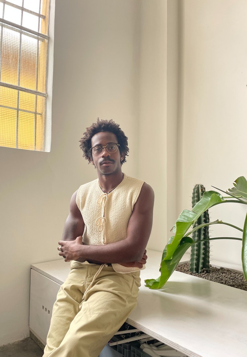 Top senza maniche beige con texture e dettaglio a fiocco, abbinato a pantaloni giallo chiaro. Sullo sfondo si vedono piante verdi e una grande finestra.