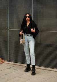 Μαύρο πουκάμισο με κουμπιά, ανοιχτό μπλε τζιν ψηλής μέσης, μαύρες μποτάκια με κορδόνια και γκρι τσάντα χιαστί. Το φόντο έχει σχέδιο από πλέγμα.