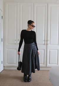 Blusa preta de mangas longas, saia maxi cinza xadrez com fivelas laterais, botas pretas de tornozelo, pequena bolsa preta, contra um fundo de armário branco.