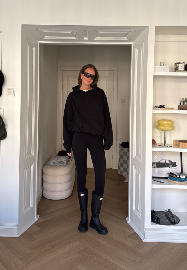 Una mujer con gafas de sol, una sudadera negra con capucha, leggings y botas hasta la rodilla está de pie en una entrada, junto a unas estanterías y artículos de decoración para el hogar.