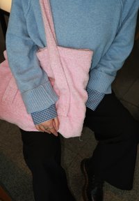 Uma bolsa de ombro rosa, fofa e de tecido, segurada por uma pessoa que veste um suéter azul claro sobre uma camisa xadrez azul, com calças pretas.