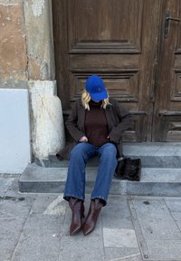 Pull en laine marron, manteau marron foncé, casquette de baseball bleue, jeans évasés, et bottines marron à bout pointu. Sac à épaule noir à proximité.