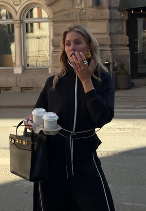 Mujer vestida de negro sostiene dos vasos de café para llevar y un bolso negro, de pie en una calle de la ciudad con un edificio de piedra ornamentado al fondo.