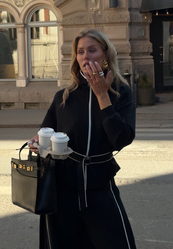 Kvinde i sort outfit holder to takeaway kaffekopper og en sort håndtaske, stående på en bygade med en udsmykket stenbygning i baggrunden.