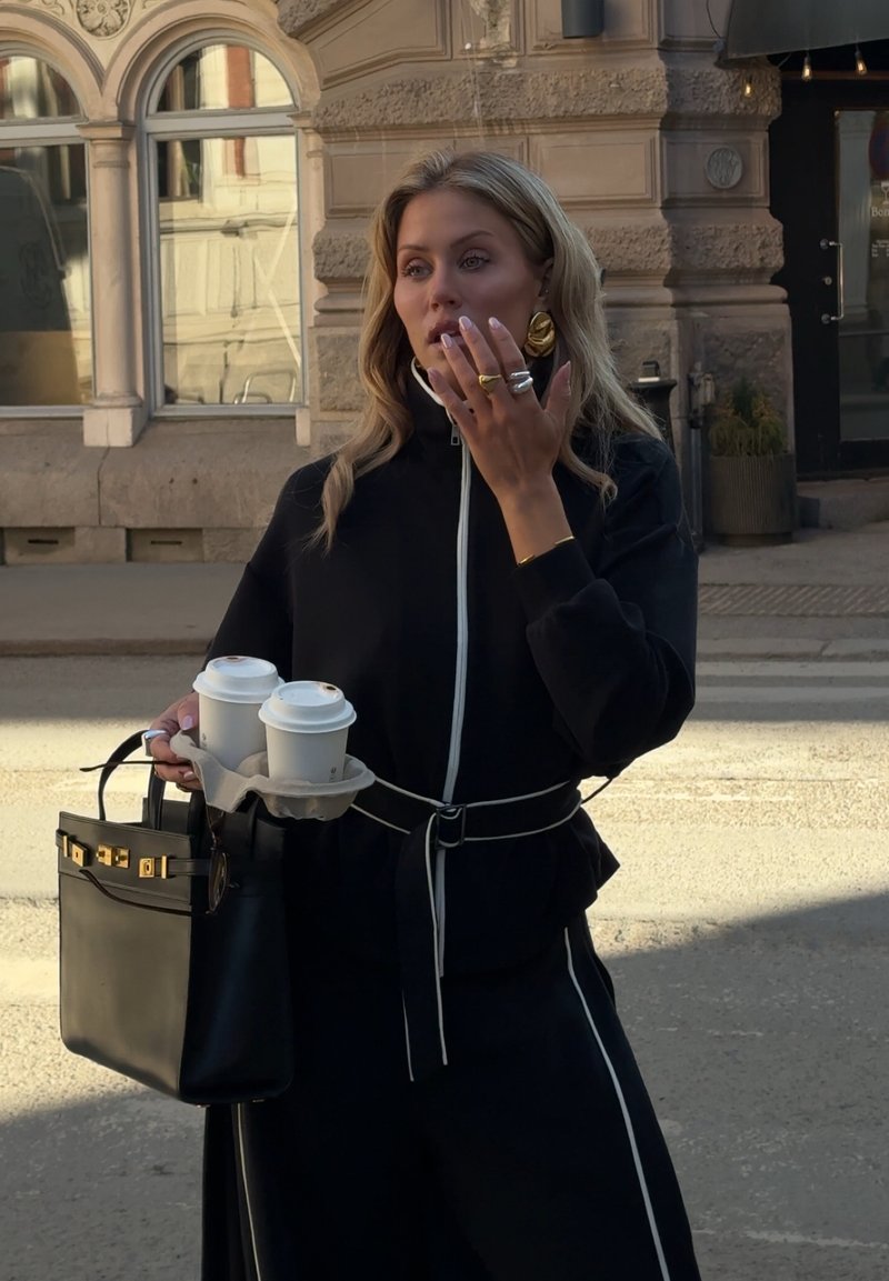 Frau in schwarzem Outfit hält zwei Kaffeebecher zum Mitnehmen und eine schwarze Handtasche, stehend auf einer Stadtstraße mit einem kunstvoll verzierten Steingebäude im Hintergrund.