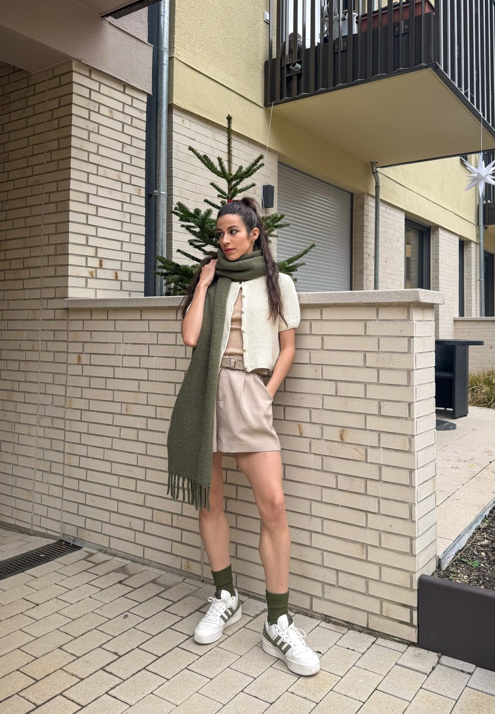 Cardigan de malha bege, lenço verde-oliva com franjas, blusa creme, calções bege, sapatilhas brancas com detalhes verdes, encostada a uma parede de tijolos.