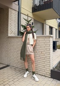 Cardigan de malha bege, lenço verde-oliva com franjas, blusa creme, calções bege, sapatilhas brancas com detalhes verdes, encostada a uma parede de tijolos.